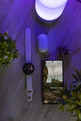 A wooden surface features a smart watch with a white band, a smartphone displaying a picturesque wallpaper of cliffs and ruins, a pair of wireless earbuds with a Samsung logo on the case, and a soft glowing light emitting a purple hue. Two small potted plants frame the scene, adding a touch of greenery.