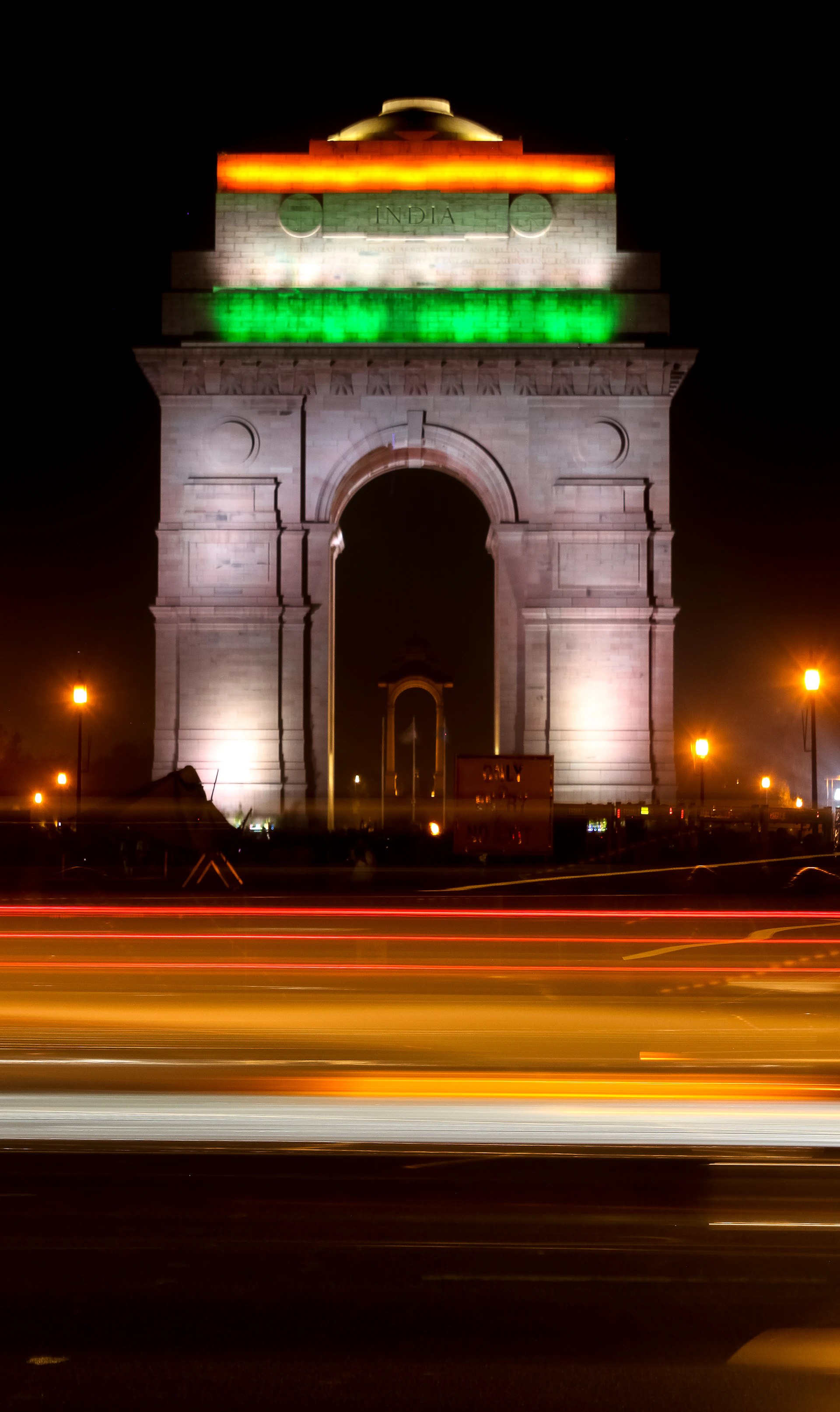 India Gate glowing warmly at sunset with a vibrant orange and purple sky in the background, framed by lush green trees.