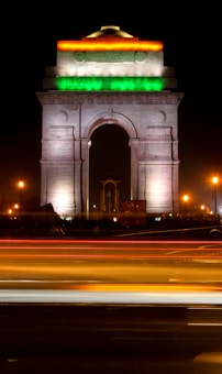 A monumental arch lit up in the colors of the Indian flag&mdash;orange, white, and green&mdash;against a dark night sky. The structure is flanked by glowing streetlights, and blurred light trails from passing vehicles create dynamic streaks across the foreground.