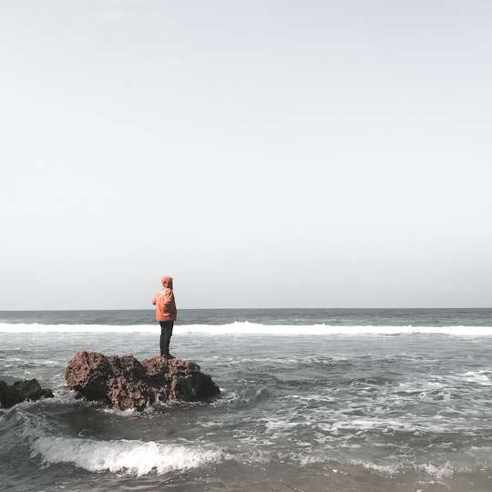 man in red jacket standing on rock in the middle of sea in Gunung-kidul Indonesia