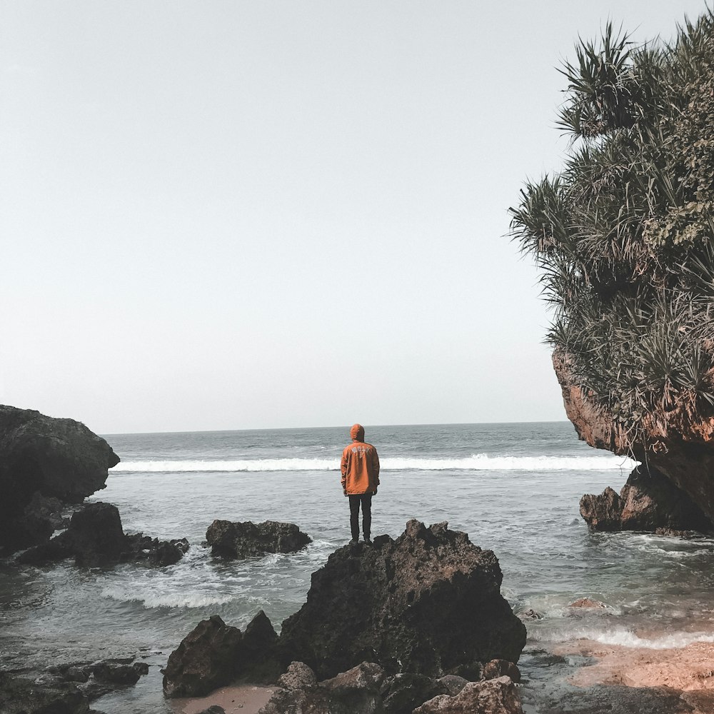 woman in orange jacket standing on rock near body of water during daytime