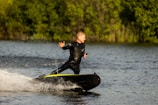 A person in a black wetsuit is riding a motorized surfboard on a body of water. They are leaning slightly back, with one arm stretched outward, creating a dynamic posture. The background consists of lush green foliage, indicating a natural and outdoor setting. Splashes of water follow the surfboard, adding movement to the scene.