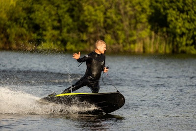 A person in a black wetsuit is riding a motorized surfboard on a body of water. They are leaning slightly back, with one arm stretched outward, creating a dynamic posture. The background consists of lush green foliage, indicating a natural and outdoor setting. Splashes of water follow the surfboard, adding movement to the scene.