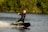 A person in a black wetsuit is riding a motorized surfboard on a body of water. They are leaning slightly back, with one arm stretched outward, creating a dynamic posture. The background consists of lush green foliage, indicating a natural and outdoor setting. Splashes of water follow the surfboard, adding movement to the scene.