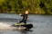 A person in a black wetsuit is riding a motorized surfboard on a body of water. They are leaning slightly back, with one arm stretched outward, creating a dynamic posture. The background consists of lush green foliage, indicating a natural and outdoor setting. Splashes of water follow the surfboard, adding movement to the scene.