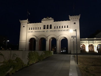 An illuminated historic building with three large arches, featuring the words 'SACHS. BAYERISCHE STAATS-EISENBAHN' across the top. Two smaller turrets flank the central structure. The scene is captured at night, with the building lights casting a warm glow. A pathway leads up to the building, bordered by greenery.