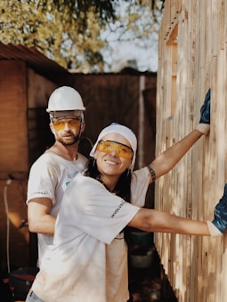 Two individuals wearing hard hats and protective eyewear are involved in construction work, assembling a wooden structure. They appear focused and collaborative, dressed casually with one of them smiling at the camera.