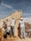 Four construction workers wearing helmets and casual work attire are assembling a wooden structure under a clear blue sky. The site is filled with construction materials and debris, with a partially constructed building visible in the background.