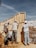 Four construction workers wearing helmets and casual work attire are assembling a wooden structure under a clear blue sky. The site is filled with construction materials and debris, with a partially constructed building visible in the background.