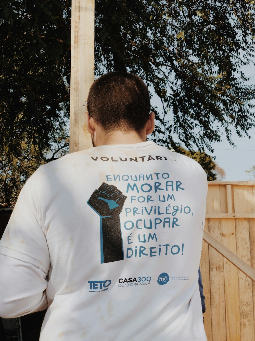 man in white and black crew neck t-shirt standing near brown wooden fence during daytime