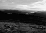 A landscape with rolling hills in the foreground, leading to a distant view of mountains. A large lake stretches across the middle of the scene, reflecting light from the overcast sky. The image is in black and white, giving it a timeless and serene quality.