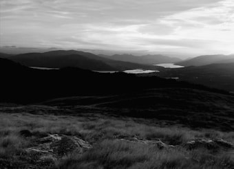 A landscape with rolling hills in the foreground, leading to a distant view of mountains. A large lake stretches across the middle of the scene, reflecting light from the overcast sky. The image is in black and white, giving it a timeless and serene quality.