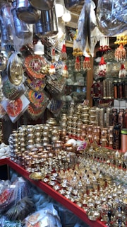 A market stall displays a variety of shiny metal items, including bowls, cups, and decorative pieces, with intricate designs. The items are made from materials like brass and copper. Decorative hangings and vibrant items are suspended above the stall, adding color and texture.