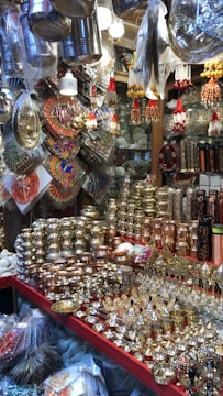 A market stall displays a variety of shiny metal items, including bowls, cups, and decorative pieces, with intricate designs. The items are made from materials like brass and copper. Decorative hangings and vibrant items are suspended above the stall, adding color and texture.