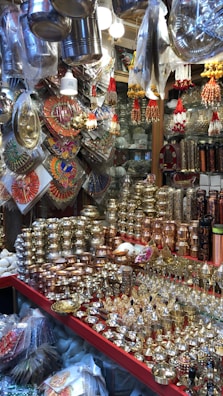 A market stall displays a variety of shiny metal items, including bowls, cups, and decorative pieces, with intricate designs. The items are made from materials like brass and copper. Decorative hangings and vibrant items are suspended above the stall, adding color and texture.