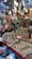 A market stall displays a variety of shiny metal items, including bowls, cups, and decorative pieces, with intricate designs. The items are made from materials like brass and copper. Decorative hangings and vibrant items are suspended above the stall, adding color and texture.