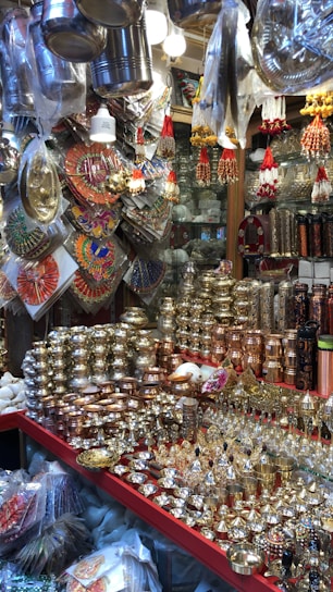 A market stall displays a variety of shiny metal items, including bowls, cups, and decorative pieces, with intricate designs. The items are made from materials like brass and copper. Decorative hangings and vibrant items are suspended above the stall, adding color and texture.