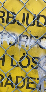 A bright yellow caution sign hanging on a chain-link fence at a construction site.