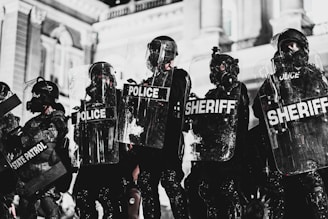 A line of law enforcement officers wearing protective gear and holding transparent shields marked with various titles such as 'Police,' 'Sheriff,' and 'State Patrol.' They are positioned in front of a grand building with detailed architecture, suggesting a tense or prepared atmosphere, possibly related to crowd control or a protest.