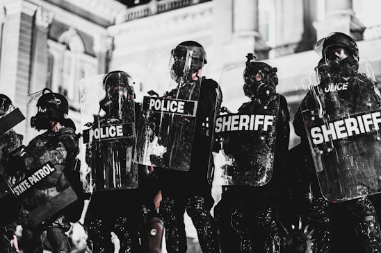 A line of law enforcement officers wearing protective gear and holding transparent shields marked with various titles such as 'Police,' 'Sheriff,' and 'State Patrol.' They are positioned in front of a grand building with detailed architecture, suggesting a tense or prepared atmosphere, possibly related to crowd control or a protest.