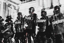A line of law enforcement officers wearing protective gear and holding transparent shields marked with various titles such as 'Police,' 'Sheriff,' and 'State Patrol.' They are positioned in front of a grand building with detailed architecture, suggesting a tense or prepared atmosphere, possibly related to crowd control or a protest.