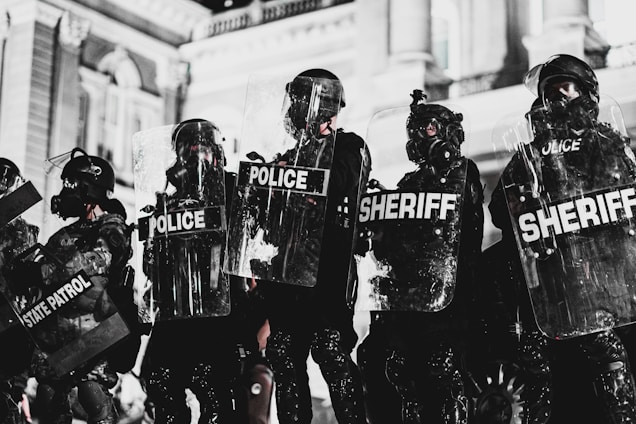 A line of law enforcement officers wearing protective gear and holding transparent shields marked with various titles such as 'Police,' 'Sheriff,' and 'State Patrol.' They are positioned in front of a grand building with detailed architecture, suggesting a tense or prepared atmosphere, possibly related to crowd control or a protest.