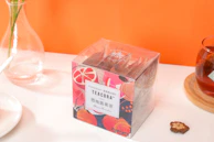 A bright and colorful tea packaging with fruit-themed designs sits on a table. A glass vase with green stems is placed nearby, alongside a cup of tea on a wooden coaster and a small decorative item resembling a dried flower. An orange background adds vibrancy to the scene.