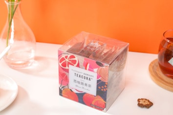 A bright and colorful tea packaging with fruit-themed designs sits on a table. A glass vase with green stems is placed nearby, alongside a cup of tea on a wooden coaster and a small decorative item resembling a dried flower. An orange background adds vibrancy to the scene.
