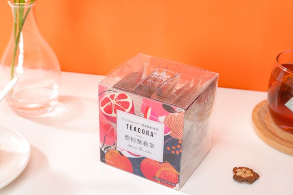 A bright and colorful tea packaging with fruit-themed designs sits on a table. A glass vase with green stems is placed nearby, alongside a cup of tea on a wooden coaster and a small decorative item resembling a dried flower. An orange background adds vibrancy to the scene.