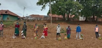A group of people standing in a line, each with a package of supplies on the ground in front of them. They are wearing protective face masks and are spread apart in an outdoor setting with green trees and traditional clay tile-roofed buildings in the background.