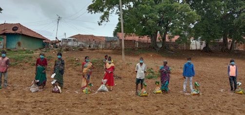A group of people standing in a line, each with a package of supplies on the ground in front of them. They are wearing protective face masks and are spread apart in an outdoor setting with green trees and traditional clay tile-roofed buildings in the background.
