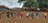 A group of people standing in a line, each with a package of supplies on the ground in front of them. They are wearing protective face masks and are spread apart in an outdoor setting with green trees and traditional clay tile-roofed buildings in the background.
