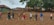 A group of people standing in a line, each with a package of supplies on the ground in front of them. They are wearing protective face masks and are spread apart in an outdoor setting with green trees and traditional clay tile-roofed buildings in the background.