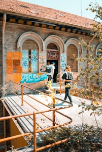 Two individuals with backpacks enter an old, abandoned building covered in colorful graffiti. The structure features arched windows and a weathered roof. Rusty metal railings and overgrown plants are visible around the walkway.
