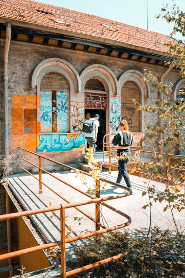 Two individuals with backpacks enter an old, abandoned building covered in colorful graffiti. The structure features arched windows and a weathered roof. Rusty metal railings and overgrown plants are visible around the walkway.