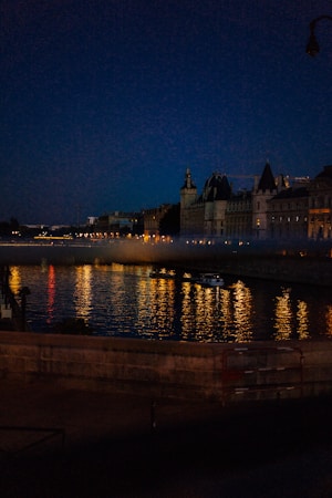 A dimly lit cityscape at night with reflections shimmering across a river. Historic buildings line the riverbank, illuminated by warm lights that create a vivid contrast against the dark sky. A boat is visible on the water, adding a serene element to the scene.