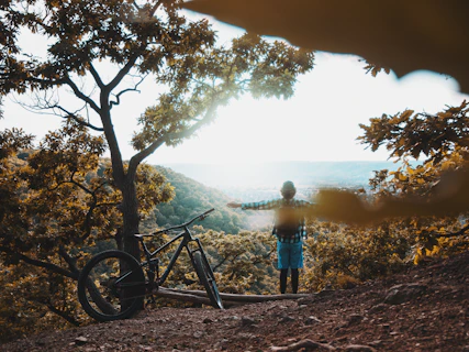 man in blue shirt standing beside black mountain bike near tree during daytime