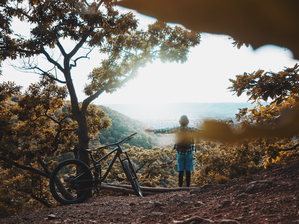 man in blue shirt standing beside black mountain bike near tree during daytime