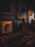 A vintage, dimly lit room with an old radio on a wooden desk, surrounded by book shelves and framed paintings. Soft, warm lighting from the lamp casts shadows, enhancing the nostalgic atmosphere.
