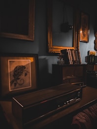 A vintage, dimly lit room with an old radio on a wooden desk, surrounded by book shelves and framed paintings. Soft, warm lighting from the lamp casts shadows, enhancing the nostalgic atmosphere.