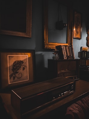 A vintage, dimly lit room with an old radio on a wooden desk, surrounded by book shelves and framed paintings. Soft, warm lighting from the lamp casts shadows, enhancing the nostalgic atmosphere.