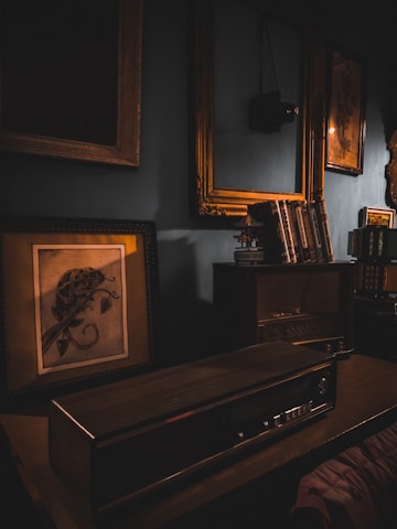 A vintage, dimly lit room with an old radio on a wooden desk, surrounded by book shelves and framed paintings. Soft, warm lighting from the lamp casts shadows, enhancing the nostalgic atmosphere.