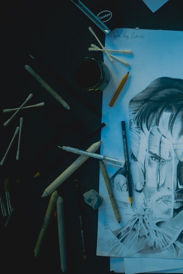 A detailed pencil drawing of a face surrounded by various art supplies including pencils, cotton swabs, and blending tools. The drawing is realistic, depicting a person with intense eyes, and is partially covered by some of the tools. The setup is on a dark surface, creating a contrast between the drawing and the background.