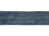 A rectangular strip of blue denim fabric with a textured pattern. The edges appear slightly frayed, and the denim has a deep blue color with a subtle variation in the weave.
