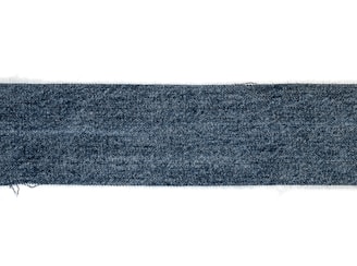 A rectangular strip of blue denim fabric with a textured pattern. The edges appear slightly frayed, and the denim has a deep blue color with a subtle variation in the weave.