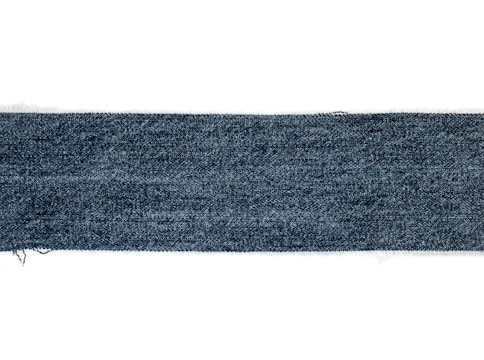 A rectangular strip of blue denim fabric with a textured pattern. The edges appear slightly frayed, and the denim has a deep blue color with a subtle variation in the weave.