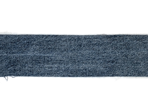A rectangular strip of blue denim fabric with a textured pattern. The edges appear slightly frayed, and the denim has a deep blue color with a subtle variation in the weave.