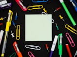 A square piece of blank paper is surrounded by various colorful office supplies including pens, paper clips, and thumbtacks, all placed on a black background. The items are arranged symmetrically around the paper, creating a neat and organized look.