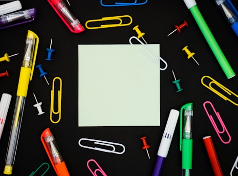 A square piece of blank paper is surrounded by various colorful office supplies including pens, paper clips, and thumbtacks, all placed on a black background. The items are arranged symmetrically around the paper, creating a neat and organized look.