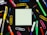 A square piece of blank paper is surrounded by various colorful office supplies including pens, paper clips, and thumbtacks, all placed on a black background. The items are arranged symmetrically around the paper, creating a neat and organized look.